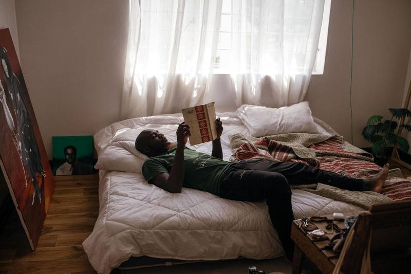 Waleed Mohammed in his bedroom in Nairobi, which doubles up as his painting studio, looking at archival material–some of the few possessions he managed to bring from Sudan.